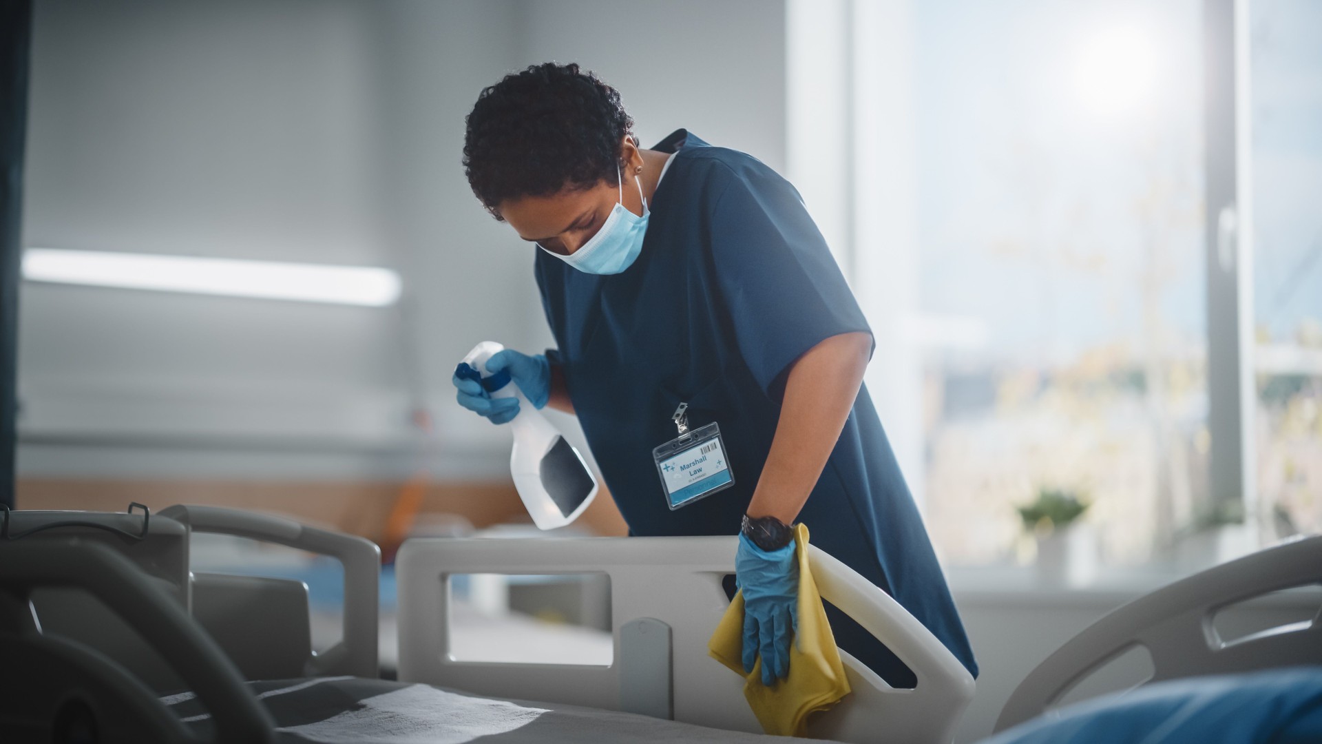 Hospital Ward: Professional Black Nurse Wearing Face Mask, Wiping the Bed, Cleaning Room After Covid-19 Patients Recover. Disinfection, Sterilizing, Sanitizing Clinic after Coronavirus Infected People